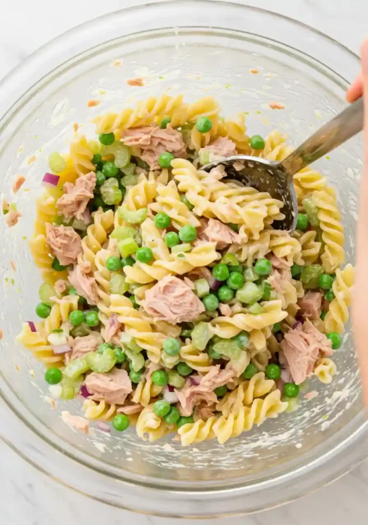 Spoon mixing pasta, tuna, celery, peas, and dressing
