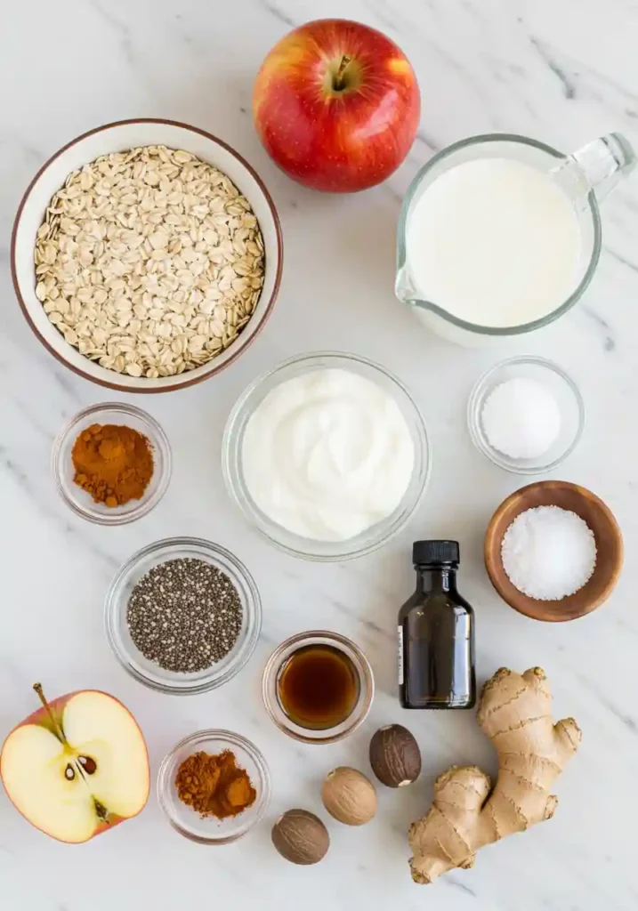 Ingredients for apple pie overnight oats including rolled oats, fresh apples, cinnamon, Greek yogurt, chia seeds, and maple syrup arranged on white background