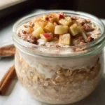 Apple pie overnight oats in a glass mason jar topped with diced apples, cinnamon, and chopped walnuts on a wooden table