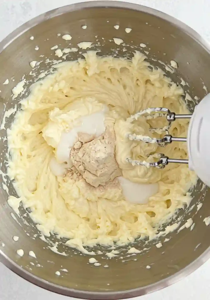 Electric mixer blending cream cheese, pudding mix, and milk in a large glass bowl