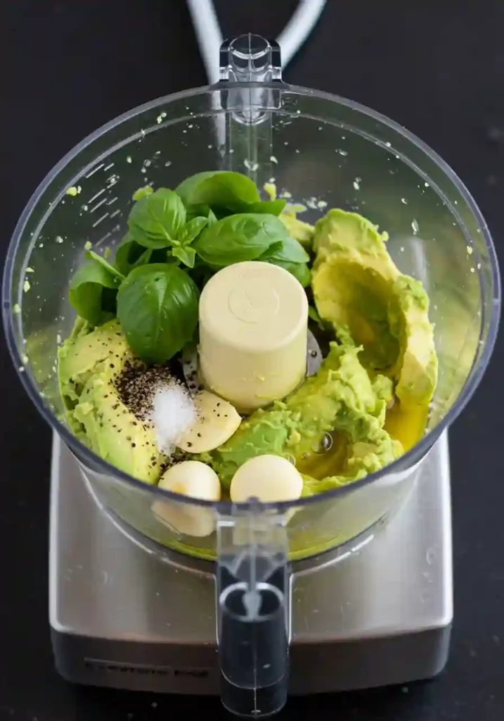 Food processor blending ripe avocados with fresh herbs, garlic, and olive oil to create smooth green sauce