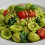 Bowl of creamy avocado pasta sauce garnished with fresh basil, cherry tomatoes, and pine nuts on a white plate