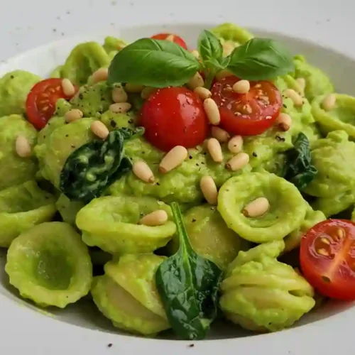 Bowl of creamy avocado pasta sauce garnished with fresh basil, cherry tomatoes, and pine nuts on a white plate