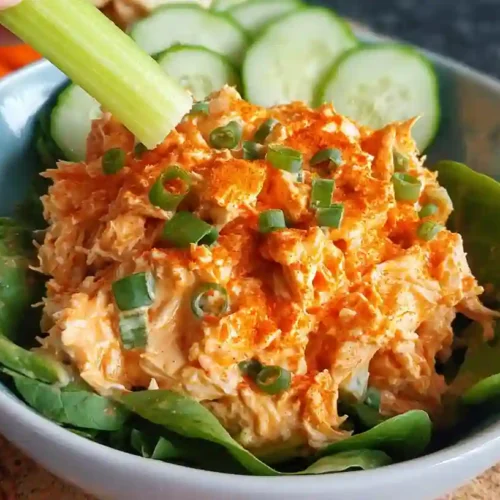 Bowl of creamy buffalo chicken salad with shredded chicken, diced celery, and red onions in a white bowl, garnished with green onions and served with crackers on a wooden cutting board