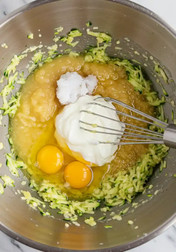 Hands whisking wet ingredients including grated zucchini, Greek yogurt, honey, and eggs in clear mixing bowl for chocolate zucchini muffins