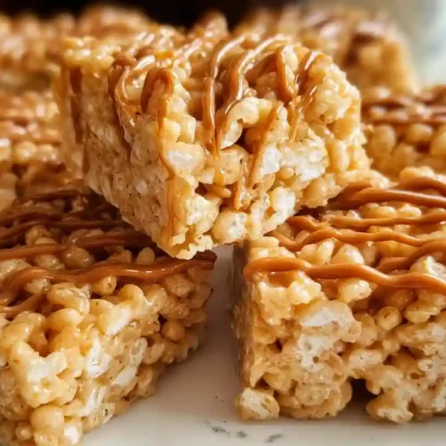 Peanut butter rice krispie treats cut into squares on a white plate with one piece lifted to show chewy texture
