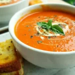 Homemade creamy tomato basil soup in white bowl with fresh basil garnish and grilled cheese sandwich